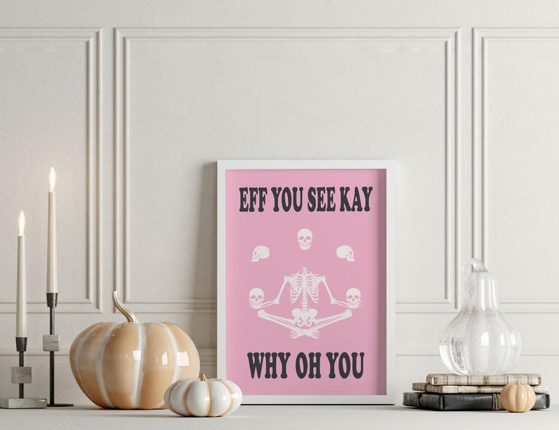 May include: A framed pink print with white text and a skeleton illustration. The text reads "EFF YOU SEE KAY WHY OH YOU". The skeleton is in a yoga pose, holding skulls. Decorative pumpkins and candles are in the foreground.