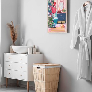 May include: A white bathroom with a white vanity, a white sink, and a wicker laundry basket. There is a framed print of a flamingo in a bathroom setting on the wall. A white bathrobe is hanging on a hook.
