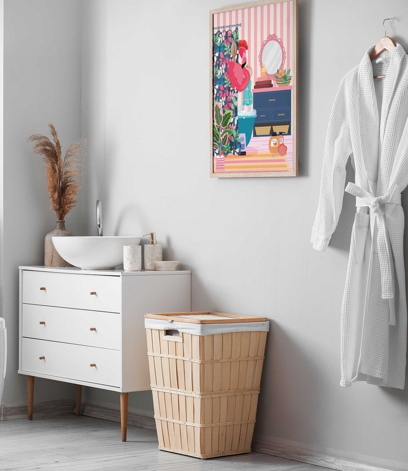 May include: A white bathroom with a white vanity, a white sink, and a wicker laundry basket. There is a framed print of a flamingo in a bathroom setting on the wall. A white bathrobe is hanging on a hook.