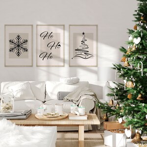 May include: Three framed prints with a white background and black designs. The prints feature a snowflake, the words "Ho Ho Ho", and a stylized Christmas tree. A white couch with a throw blanket and pillows sits in front of the prints. A Christmas tree with gold ornaments and white lights is in the corner of the room.