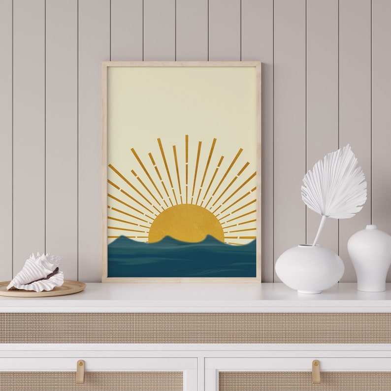 May include: A framed print with a minimalist design of a yellow sun rising over blue waves. The sun has a stylized, geometric shape with rays extending outward. The background is a light yellow.