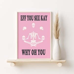 May include: Pink framed art print with a white skeleton in a yoga pose, surrounded by skulls. The text reads "EFF YOU SEE KAY WHY OH YOU" in black. The artwork is displayed on a wooden shelf.