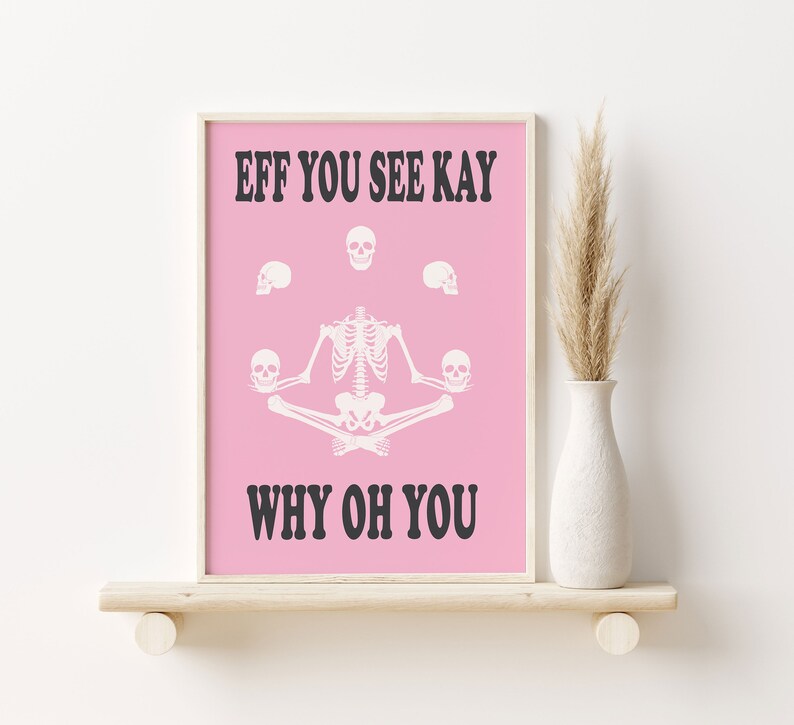 May include: Pink framed art print with a white skeleton in a yoga pose, surrounded by skulls. The text reads "EFF YOU SEE KAY WHY OH YOU" in black. The artwork is displayed on a wooden shelf.