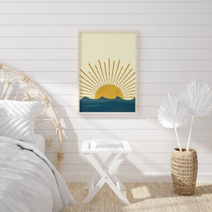 May include: A framed print of a minimalist sun rising over a blue mountain range. The sun is yellow and the mountains are a deep blue. The print is hanging on a white shiplap wall.