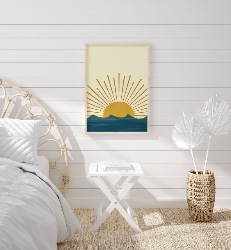 May include: A framed print of a minimalist sun rising over a blue mountain range. The sun is yellow and the mountains are a deep blue. The print is hanging on a white shiplap wall.