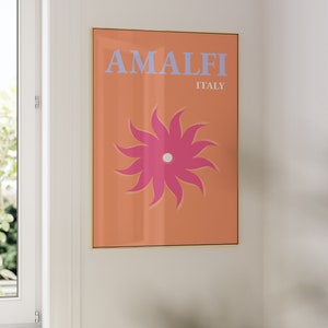 K&ouml;nnte beinhalten: Ein gerahmter Druck mit einem pfirsichfarbenen Hintergrund und einer stilisierten rosa Blume mit wei&szlig;em Zentrum. Der Text "AMALFI ITALIEN" ist in hellblau gedruckt.
