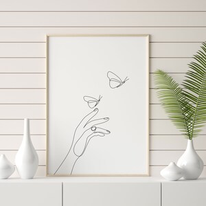May include: A black and white line art print of a hand reaching out to two butterflies. The print is framed in a light wood frame and is hanging on a white wall.