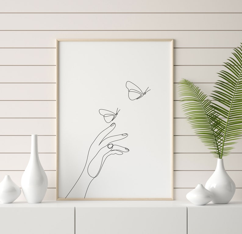 May include: A black and white line art print of a hand reaching out to two butterflies. The print is framed in a light wood frame and is hanging on a white wall.
