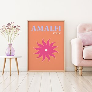 K&ouml;nnte beinhalten: Ein pfirsichfarbener Poster mit dem Text "AMALFI ITALIEN" in hellblau. Eine stilisierte rosa Blume mit wei&szlig;em Zentrum befindet sich in der Mitte des Posters.