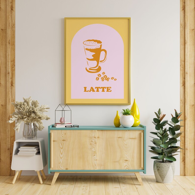 May include: A framed print with a yellow border and a pink background. The print features a stylized illustration of a latte in a glass with a brown handle. The text "LATTE" is printed in orange below the illustration.