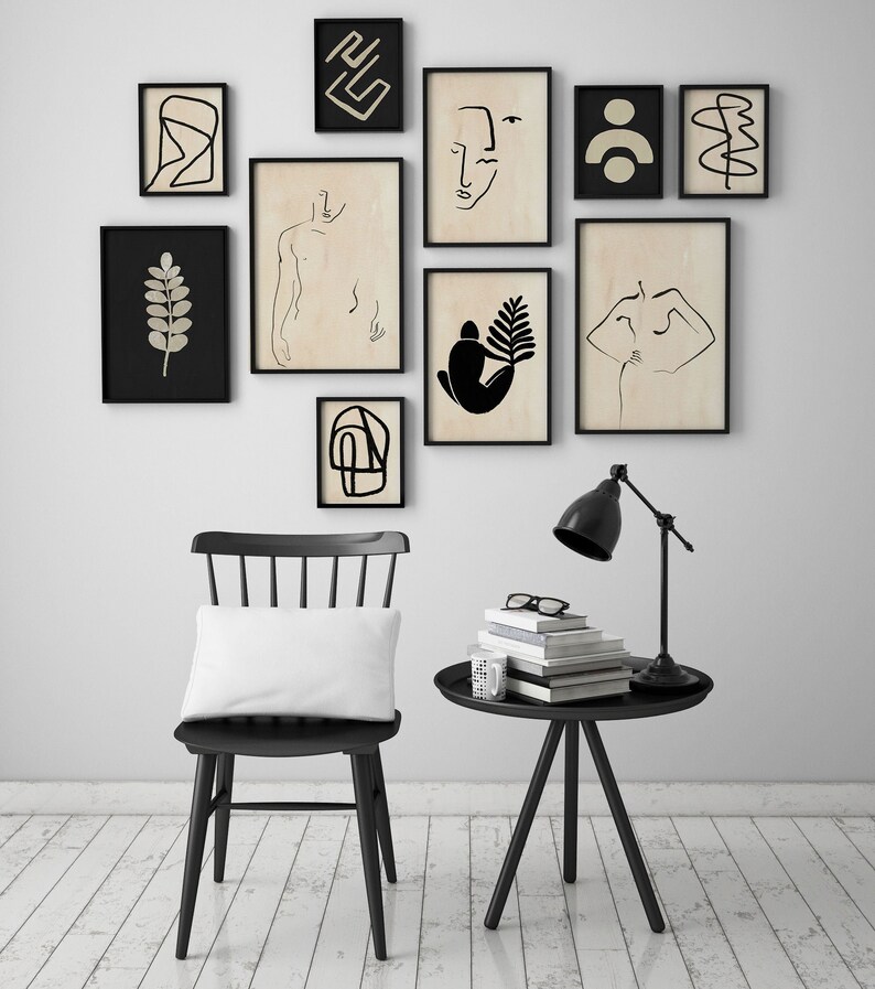 May include: A black chair with a white pillow sits in front of a white wall with black framed artwork. A black side table with a lamp and a stack of books sits next to the chair.