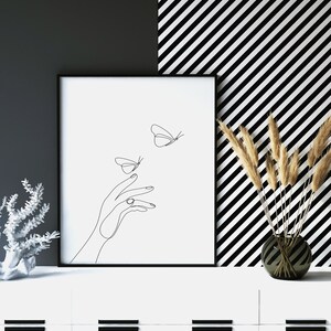 May include: Black and white line art print of a hand reaching out to two butterflies. The print is framed in a black frame and is hanging on a white wall with a black and white striped background.