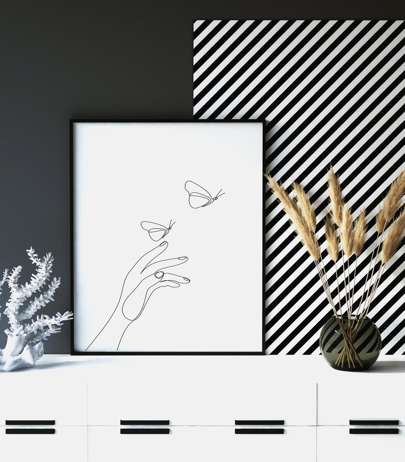 May include: Black and white line art print of a hand reaching out to two butterflies. The print is framed in a black frame and is hanging on a white wall with a black and white striped background.