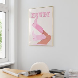 Pu&ograve; includere: Una stampa incorniciata con uno sfondo rosa e la parola "HOWDY" in lettere rosa. La stampa presenta un paio di stivali da cowboy rosa con una gamba abbronzata.