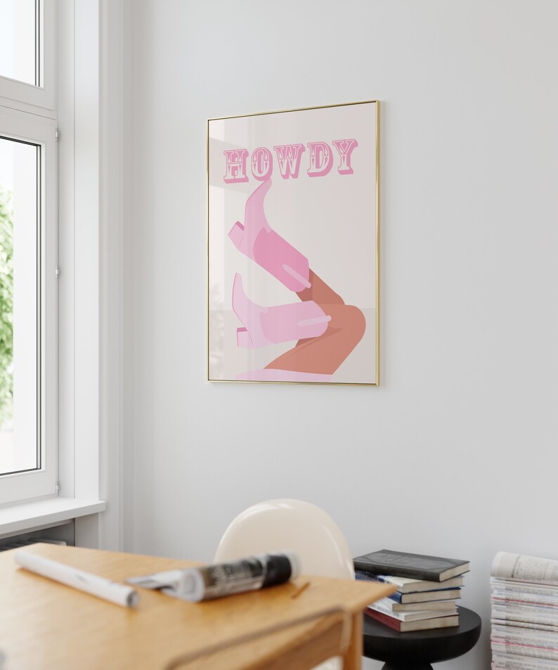 Pu&ograve; includere: Una stampa incorniciata con uno sfondo rosa e la parola "HOWDY" in lettere rosa. La stampa presenta un paio di stivali da cowboy rosa con una gamba abbronzata.