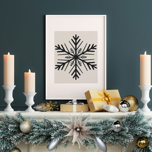 May include: A white fireplace mantel decorated with a green garland, gold and silver ornaments, and two white candle holders with lit candles. A framed print with a black snowflake design hangs above the mantel.