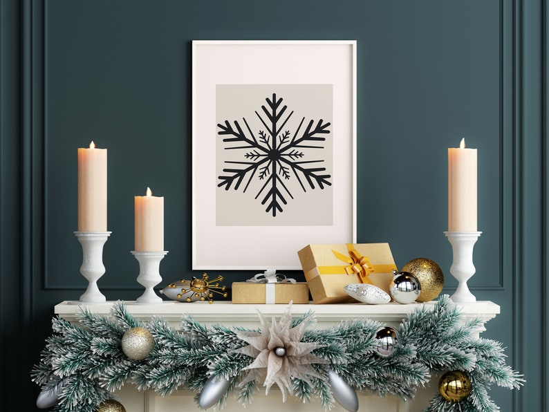 May include: A white fireplace mantel decorated with a green garland, gold and silver ornaments, and two white candle holders with lit candles. A framed print with a black snowflake design hangs above the mantel.