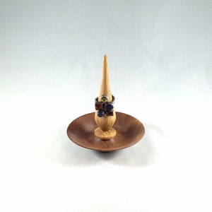 May include: A wooden ring holder with a small dish and a pointed spire. The holder is made of dark wood and has a natural finish. Several rings with colorful gemstones are displayed on the spire.