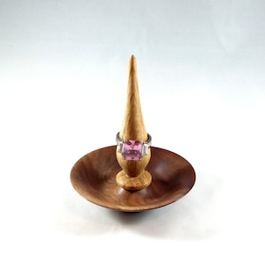 May include: A wooden ring holder with a small dish and a pointed spire. A ring with a large pink square stone and two smaller clear stones is displayed on the spire.