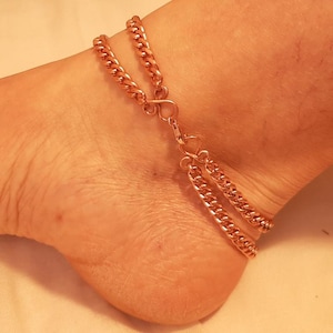 May include: A copper-coloured anklet with a double chain design. The anklet features a delicate chain that wraps around the ankle, connected by a small clasp. The chains are made of small, interlocking links.