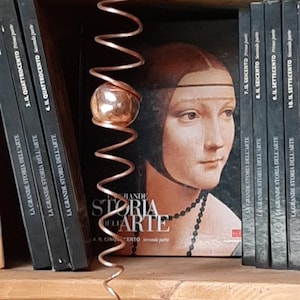 May include: A copper-coloured, spiral-shaped decorative object with a spherical element hangs in front of a shelf. Behind it are several books with the title "LA GRANDE STORIA DELL'ARTE" and a portrait of a woman.