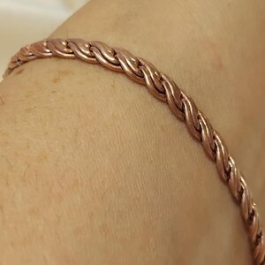 Peut inclure: Un bracelet de couleur cuivrée avec un motif tressé. Le bracelet est porté sur le bras d'une personne. Le motif tressé est constitué de brins de métal entrelacés, créant une surface texturée. Le bracelet est de couleur brun-rouge chaud.