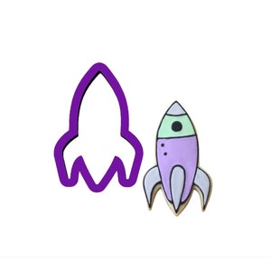 May include: A purple and green rocket cookie cutter with a white outline. The cutter is next to a decorated cookie in the shape of a rocket.