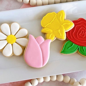 May include: A collection of four flower-shaped cookies on a white rectangular plate. The cookies include a daisy with white petals and a yellow center, a pink tulip, a yellow daffodil, and a red rose with green leaves. The cookies are decorated with icing.