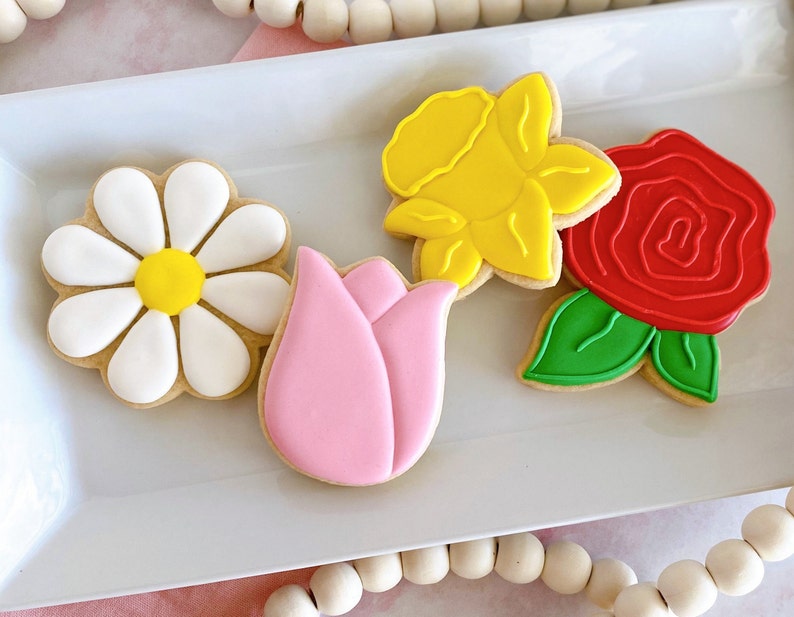 May include: A collection of four flower-shaped cookies on a white rectangular plate. The cookies include a daisy with white petals and a yellow center, a pink tulip, a yellow daffodil, and a red rose with green leaves. The cookies are decorated with icing.