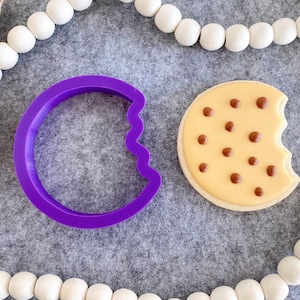 May include: Purple plastic cookie cutter shaped like a half moon with a wavy edge, next to a decorated sugar cookie with brown chocolate chips and a bite taken out.