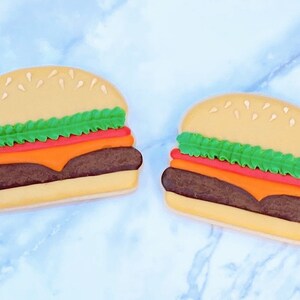 May include: Two burger-shaped cookies with sesame seed buns, green lettuce, red tomato, orange cheese, and a brown patty. The cookies are on a light blue marble surface. The cookies are a fun food-themed treat.