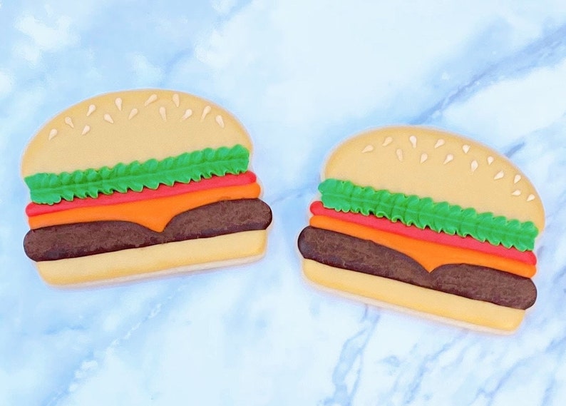 May include: Two burger-shaped cookies with sesame seed buns, green lettuce, red tomato, orange cheese, and a brown patty. The cookies are on a light blue marble surface. The cookies are a fun food-themed treat.