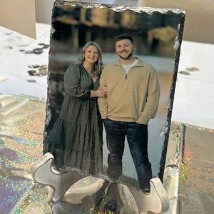 May include: A rectangular slate photo print featuring a couple, supported by a decorative metal stand. The photo shows a man in a tan sweater and jeans, and a woman in a green dress. The slate has a natural, rough edge.