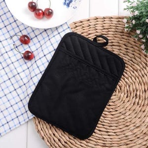 May include: A black quilted oven mitt with a pocket, featuring a loop for hanging. The mitt is on a woven placemat, with a checkered cloth and a plate of cherries in the background. The mitt is designed for kitchen use.