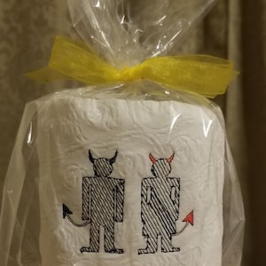 May include: White toilet paper roll with an embroidered design of two devil figures, one with black horns and the other with red horns. The roll is wrapped in clear cellophane and tied with a yellow ribbon.