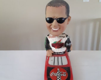 May include: A red bobblehead figurine of a race car driver wearing sunglasses and a Coca-Cola racing uniform. The driver is holding a Coca-Cola bottle and is sitting in a red race car with the Coca-Cola logo on the hood. The car has the text "The Coca-Cola Racing Family" on the bottom.