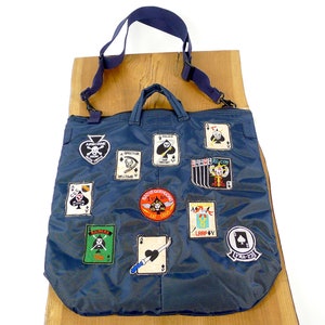 May include: A dark blue canvas tote bag with a variety of patches, including a skull and crossbones, playing cards, and military insignia. The bag has a long shoulder strap and two top handles.