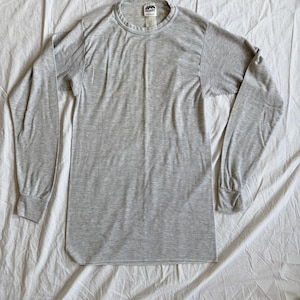May include: A light gray long-sleeve shirt with a crew neck and ribbed cuffs. The shirt is made of a lightweight material and has a tag at the neckline. The shirt is laid flat on a white surface.