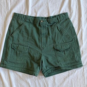 May include: Green cargo shorts with two front pockets and a button closure. The shorts are made of a durable fabric and have a classic, casual style. The shorts are a solid green color.