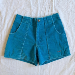 May include: Teal corduroy shorts with front pockets and a button closure. The shorts have a small red embroidered detail near the hem. The fabric has a ribbed texture. The shorts are laid flat on a white surface.