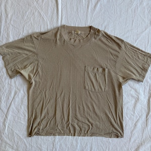 May include: A tan-colored t-shirt with a crew neck and a single pocket on the left chest. The short-sleeved shirt is made of a soft material and is laid flat on a white surface.