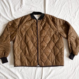 May include: Brown quilted jacket with a diamond pattern. The jacket has a black collar and cuffs. The jacket is laid out on a white surface. The jacket has a zipper closure.
