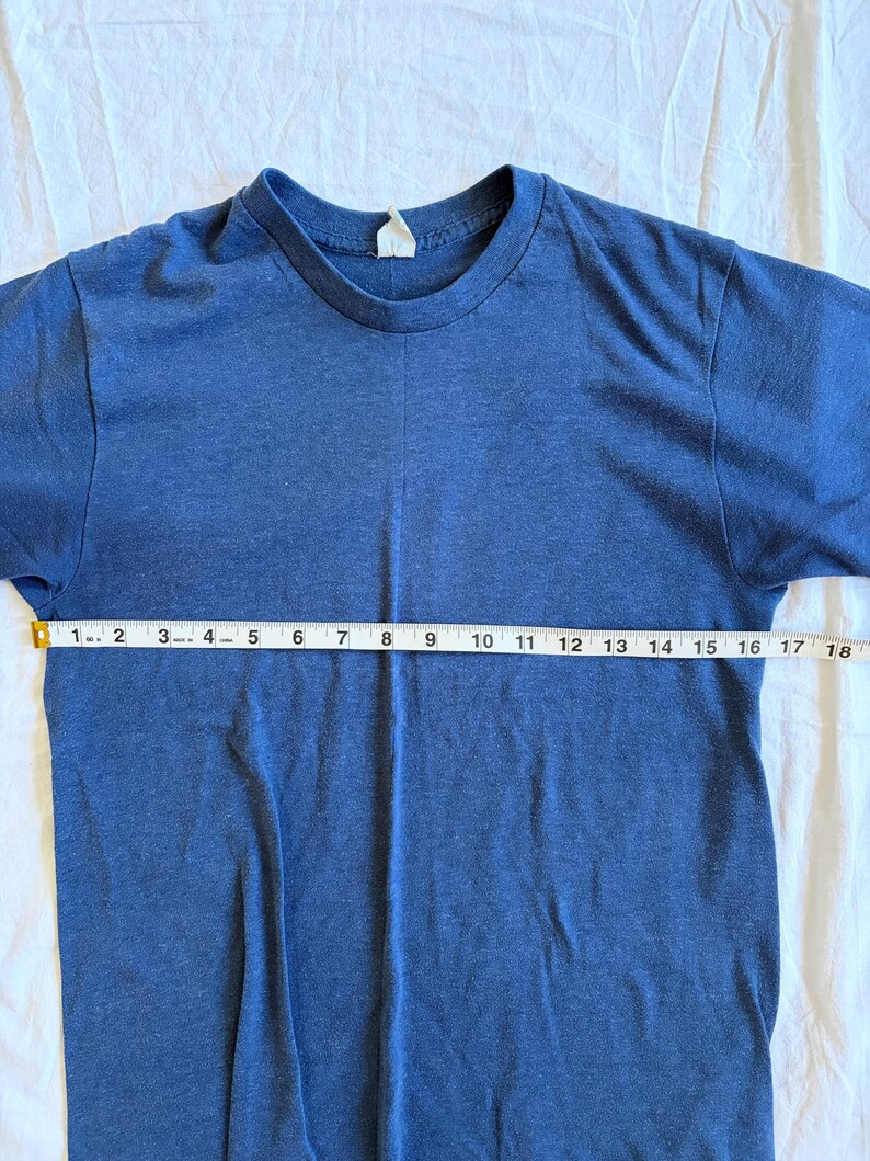 May include: A blue t-shirt with a crew neck. The shirt is measured with a tape measure, showing a length of approximately 18 inches. The fabric appears to be a soft, knit material.