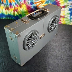 May include: A gray suitcase-style speaker with two large speakers on the front. The speakers have a black and silver design. The suitcase has a black handle and gold accents.