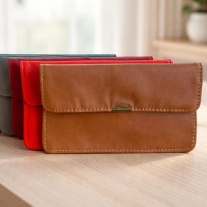 Puede incluir: Una fila de estuches de cuero en varios colores, incluyendo azul marino, verde azulado, gris, rojo y marrón. El estuche marrón está en primer plano, con un cierre de solapa y un broche metálico. Los estuches están sobre una superficie de madera.