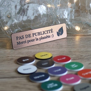 Könnte beinhalten: Ein Holzschild mit dem Text "PAS DE PUBLICITÉ Merci pour la planète :)" in schwarzen Buchstaben. Das Schild liegt auf einer Holzoberfläche mit einer Vielzahl kleiner, runder, bunter Scheiben, die um es herum verstreut sind.