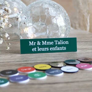 Könnte beinhalten: Ein grünes Schild mit weißem Text, auf dem "Mr & Mme Talion et leurs enfants" steht. Das Schild steht auf einem Holztisch mit verschiedenen bunten runden Gegenständen, die um es herum verstreut sind.