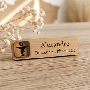 Könnte beinhalten: Ein rechteckiges Namensschild aus Holz mit einem quadratischen Metalleinsatz, der das schwarze Apothekensymbol aus Schlange und Kelch zeigt. Der Text Alexandre und Docteur en Pharmacie ist schwarz in das helle Holz eingraviert.