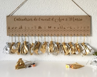 Puede incluir: Un calendario de adviento de madera con 25 casillas numeradas, cada una con una pequeña bolsa dorada o plateada colgando. El calendario está personalizado con el texto "Calendrier de l'avent d' Ava & Marin".