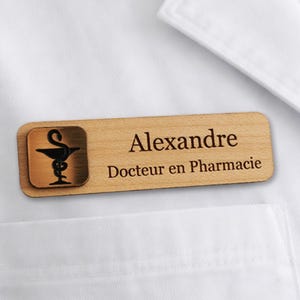 Peut inclure: Un badge en bois avec le nom "Alexandre" et le titre "Docteur en Pharmacie" gravés en brun foncé. Le badge comporte un carré de couleur bronze avec un symbole de pharmacie. Le badge est épinglé sur un vêtement blanc.